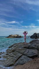 A woman raising her arms on a rocky coast at Playa Cala Sa Boadella, Spain. Slim silhouette, summer light, perfect for motivational or wellness poster.