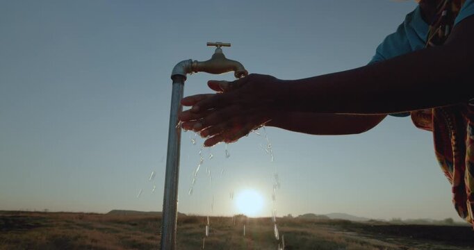 Backlit cropped close-up of a Black African woman in traditional clothing washing her hands at a faucet at sunset