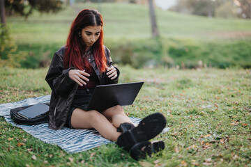 A young girl with red hair uses a laptop on a blanket in nature, surrounded by grass.