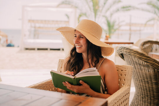 A woman enjoys reading a book at a beachside caf, wearing a large sun hat - Powered by Adobe