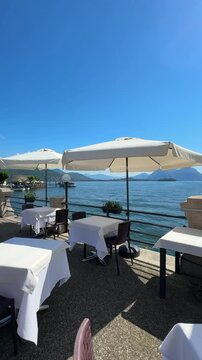 Street caf&eacute; with white tablecloths and sun umbrellas on the Lake Maggiore promenade. Summer vibe, waterfront dining, and elegant Italian ambiance in the lakeside town