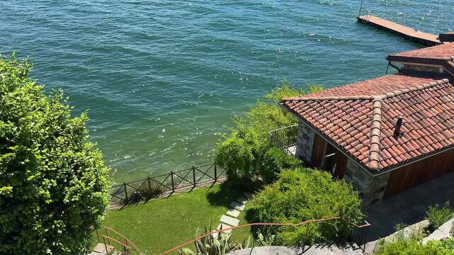 Tranquil view of a charming courtyard near Lake Como with a traditional tiled-roof house, a stone path, and a small pier for water access or docking a boat. Peaceful Italian lakeside elegance
