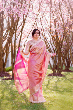 A woman in a shimmering pink traditional outfit stands serenely among cherry blossoms, radiating warmth, elegance, and cultural pride.