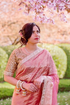 A woman in a shimmering pink traditional outfit stands serenely among cherry blossoms, radiating warmth, elegance, and cultural pride.