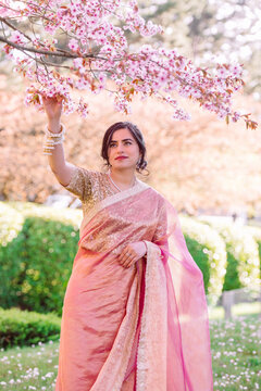 A woman in a shimmering pink traditional outfit stands serenely among cherry blossoms, radiating warmth, elegance, and cultural pride.