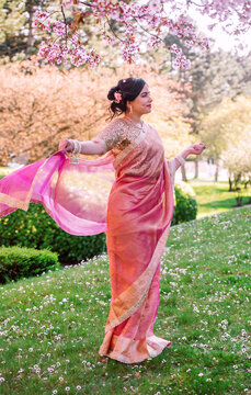 A woman in a shimmering pink traditional outfit stands serenely among cherry blossoms, radiating warmth, elegance, and cultural pride.