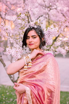 A woman in a shimmering pink traditional outfit stands serenely among cherry blossoms, radiating warmth, elegance, and cultural pride.