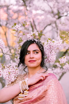 A woman in a shimmering pink traditional outfit stands serenely among cherry blossoms, radiating warmth, elegance, and cultural pride.