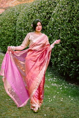 A woman in a shimmering pink traditional outfit stands serenely among cherry blossoms, radiating warmth, elegance, and cultural pride.