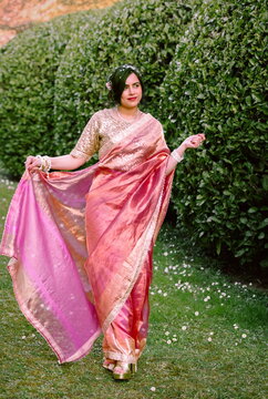 A woman in a shimmering pink traditional outfit stands serenely among cherry blossoms, radiating warmth, elegance, and cultural pride.