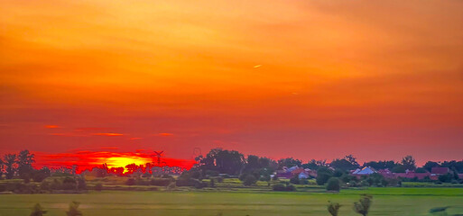 Obraz premium Dramatic sunset over a rural landscape with trees, fields, and colorful sky. Vibrant orange and red tones create a peaceful and dreamy countryside evening atmosphere.