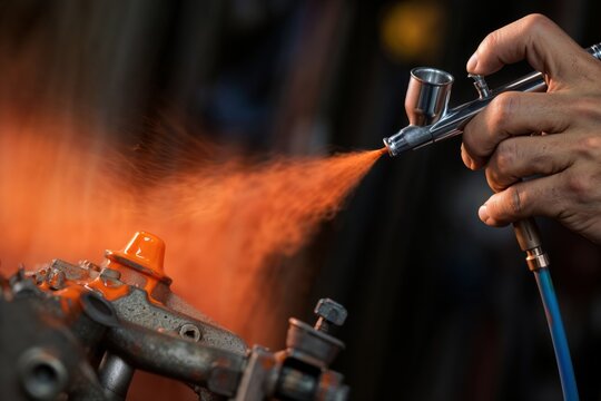 Airbrushing a metal part. A professional's hand holds an airbrush, spraying a vibrant orange coating onto a custom automotive or industrial machine part.