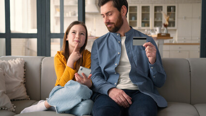Joyful Caucasian parent man father holding credit card guy male talking finance smiling little schoolgirl girl child using mobile phone e-commerce at home family remote kid daughter shopping dad pay