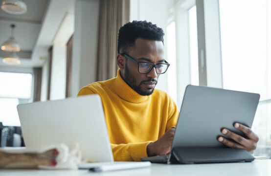 African American businessman works on laptop, tablet in bright office space. Focused pro wears yellow turtleneck sweater, glasses, listening to music via earphones. Tech startup environment for web