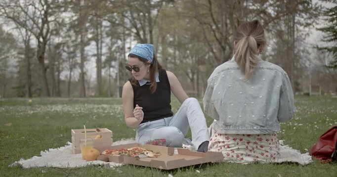 Two young women enjoy a relaxing picnic in the park with pizza, fruit, and drinks. They talk and have fun, spending quality time together on a beautiful day.
