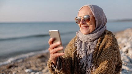 Senior woman enjoys beach, video calling with device, wearing shawl and sunglasses. Relaxing seaside connection, capturing coastal moments.