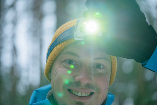 Hiker wearing a yellow beanie and blue jacket smiling and pointing a headlamp in a winter forest at night, enjoying the adventure and exploring nature