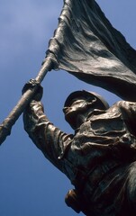 Fototapeta premium A monument of an Asian soldier holding up the flag, against a blue sky background, shot from a low angle with a wide-angle lens, in a sculptural style with bronze material, displaying a strong contras