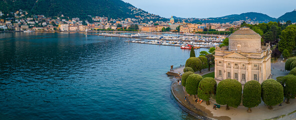 Fototapeta premium Aerial view of Volta Temple in Como, Italy, surrounded by park trees, lakefront promenade, and clear blue lake under sunny summer skies
