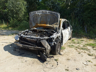 A charred and gutted vehicle stands abandoned on a dirt path, its hood open revealing the burnt remains of the engine.