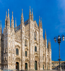Gothic-style Milan Cathedral facade with ornate spires and intricate stonework against clear blue sky, captured in daylight