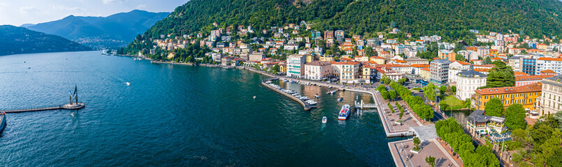 Aerial summer view of Lake Como with deep blue water, surrounding green hills, villas, and boats under a clear, sunny sky in Italy