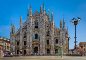 Gothic-style Milan Cathedral facade with ornate spires and intricate stonework against clear blue...