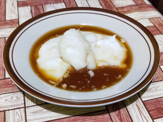 Bubur sumsum, traditional Indonesian food. Soft white marrow porridge served on a plate sprinkled with thick brown sugar sauce on a wooden patterned table.