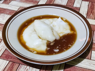 Bubur sumsum, traditional Indonesian food. Soft white marrow porridge served on a plate sprinkled with thick brown sugar sauce on a wooden patterned table.