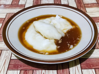 Bubur sumsum, traditional Indonesian food. Soft white marrow porridge served on a plate sprinkled with thick brown sugar sauce on a wooden patterned table.