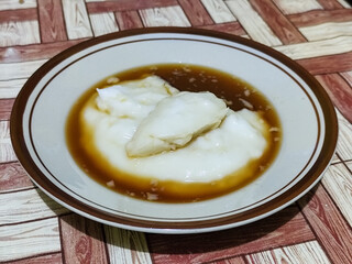 Bubur sumsum, traditional Indonesian food. Soft white marrow porridge served on a plate sprinkled with thick brown sugar sauce on a wooden patterned table.