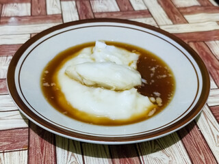 Bubur sumsum, traditional Indonesian food. Soft white marrow porridge served on a plate sprinkled with thick brown sugar sauce on a wooden patterned table.