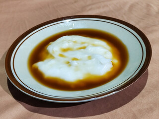 Bubur sumsum, traditional Indonesian food. Soft white marrow porridge served on a plate sprinkled with thick brown sugar sauce on a wooden patterned table.