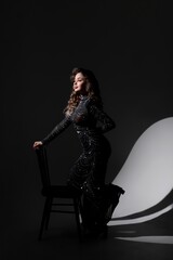 Girl in long black dress near chair in studio on black background, she has curly hair.
