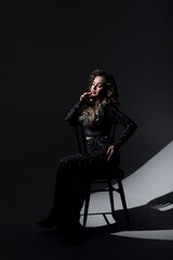 Girl in long black dress near chair in studio on black background, she has curly hair.