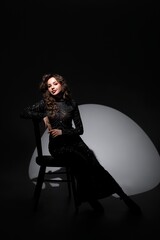 A girl in a long black dress stands near a black chair