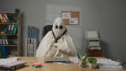 Halloween businessman ghost in white sheet sits at office desk finishes work on laptop, closes...