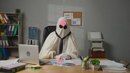 Halloween businessman ghost in white sheet sits at office desk draws smiling face on sticky note...