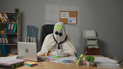 Halloween businessman ghost in white sheet sits at office desk draws sad face on sticky note and...