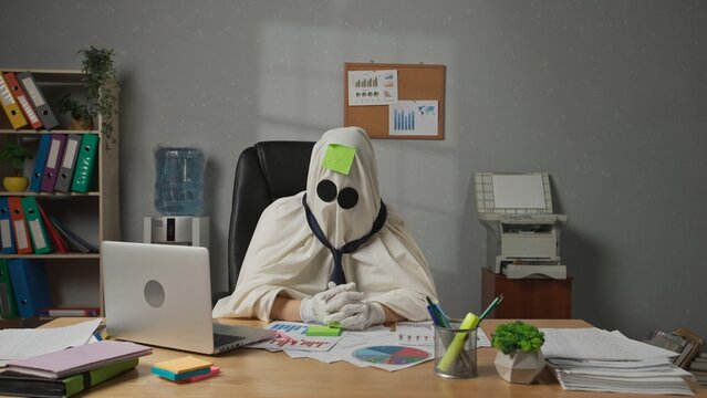 Halloween businessman ghost in white sheet sits at office desk draws sad face on sticky note and sticks it on forehead. Happy halloween business concept.
