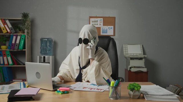 Halloween businessman ghost in white sheet with necktie sitting at office desk working on laptop, talking on smartphone. Happy halloween business concept.
