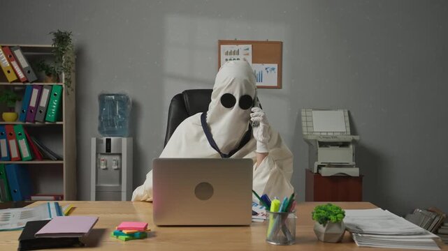 Halloween businessman ghost in white sheet with necktie sitting at office desk working on laptop, talking on smartphone. Happy halloween business concept.