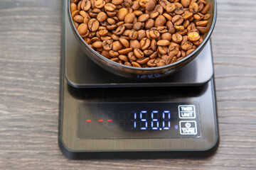 A digital scale holding a glass bowl of roasted coffee beans at 156g on a wooden table, showcasing...