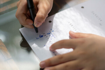Child hands focused on solving math problems with a pen on a piece of paper. The scene captures a quiet moment of concentration and learning at a reflective glass table