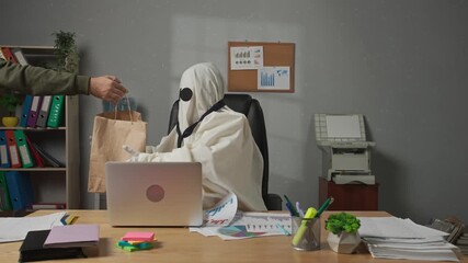 Halloween businessman ghost in white sheet sits at office desk and works, receives food delivery service paper bag with lunch. Happy halloween business concept.