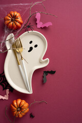 A ghost-shaped vertical plate with golden cutlery, surrounded by pumpkins, bats, and cobweb décor,...