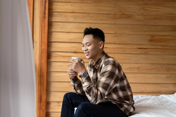 Fototapeta premium A young man enjoys a warm beverage while sitting in a bright wooden room. The cozy atmosphere reflects comfort and relaxation, making it a perfect moment for calm contemplation or a peaceful morning.