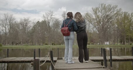 Two women enjoy a leisurely stroll on a park pier, relishing the peace and bonding time together. - Powered by Adobe