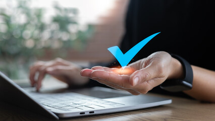 Blue check mark icon floating above a hand with laptop, symbolizing online approval, digital...