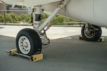 Detailed view of a military aircraft's landing gear with wheels secured by chocks. The metal structure and tire textures are clearly visible on the concrete ground.